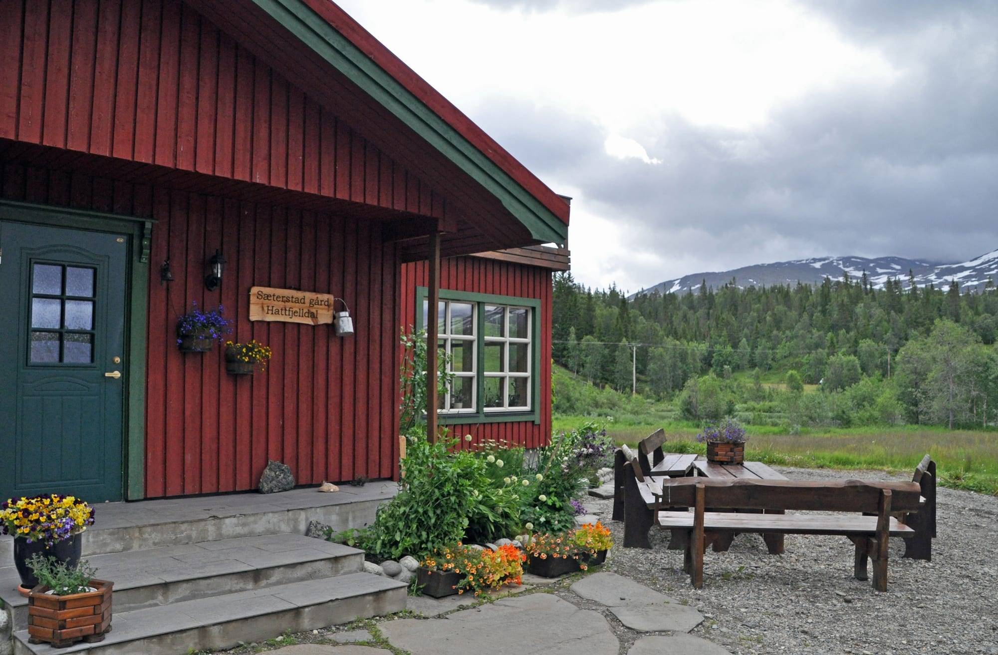 Rødt gårdshus med blomster og benk på tunet, med fjell i bakgrunnen